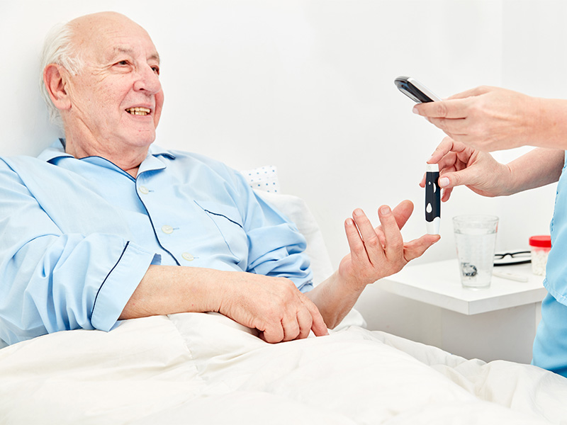 Pflegekraft misst im Schlafzimmer den Blutzucker eines Diabetikers – professionelle häusliche Krankenpflege in Ebstorf.